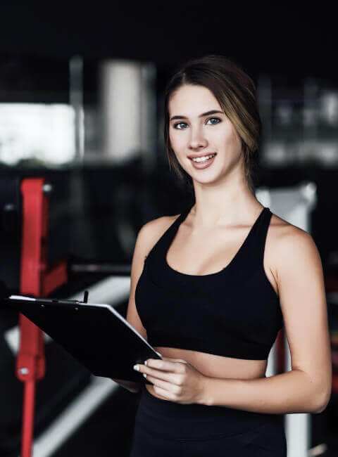 Mujer sonriente en un gimnasio sosteniendo una tablet y vistiendo ropa deportiva negra, lista para entrenar o dar instrucciones.