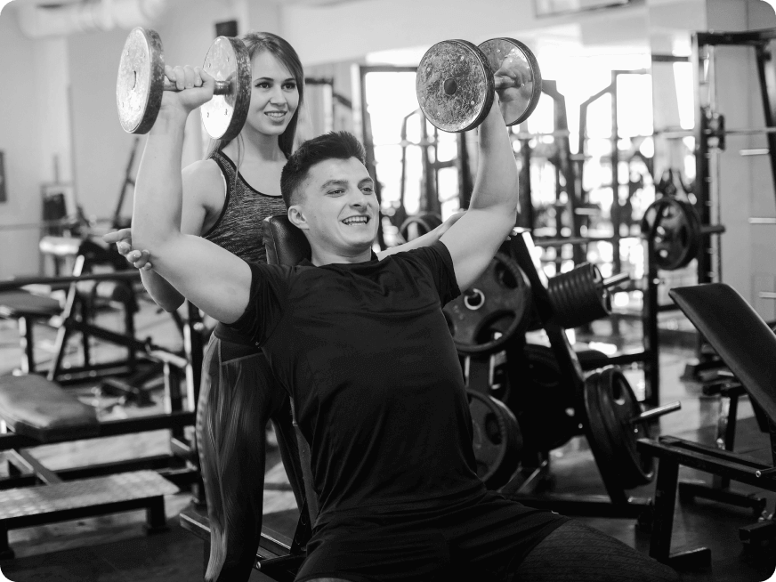 Hombre levantando pesas en un gimnasio mientras recibe apoyo de una mujer. La imagen muestra un ambiente de entrenamiento físico y motivación.