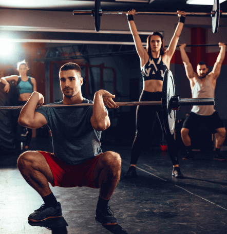 Grupo de personas entrenando en un gimnasio, con un hombre realizando una sentadilla con barra y dos mujeres levantando pesas sobre sus cabezas. Ambiente de fitness y entrenamiento de fuerza.