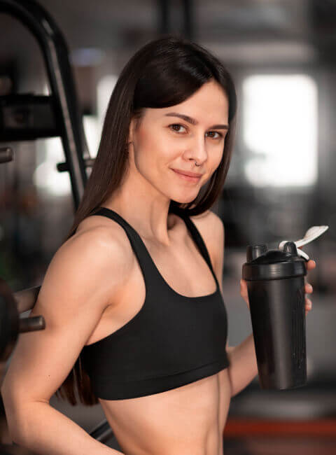 Mujer joven en un gimnasio sosteniendo un shaker negro, vistiendo un top deportivo negro, con una expresión sonriente y un fondo desenfocado de equipo de entrenamiento.