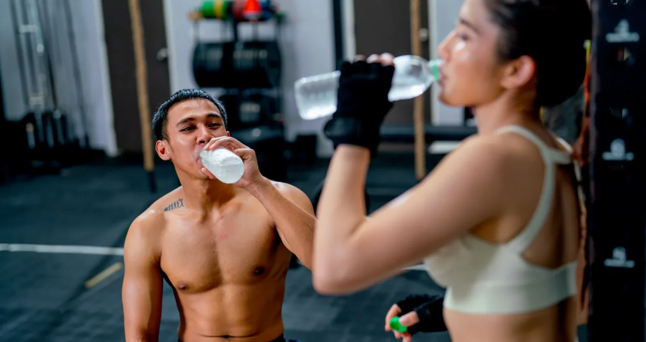Hombres y mujeres en un gimnasio bebiendo agua para mantenerse hidratados durante el entrenamiento. Ambos lucen ropa deportiva y están enfocados en su rutina de ejercicio.