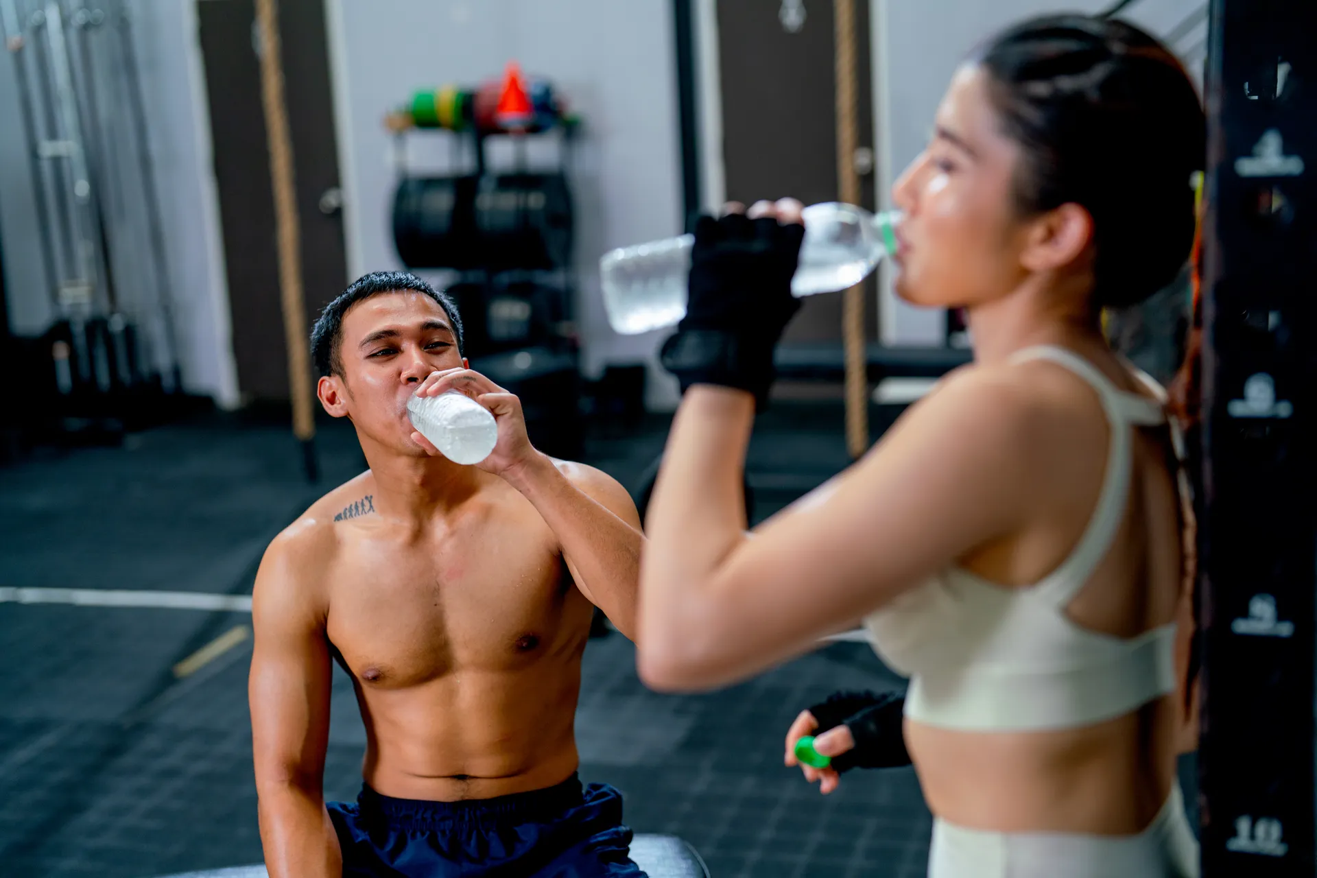 Hombres y mujeres en un gimnasio bebiendo agua para mantenerse hidratados durante el entrenamiento. Ambos lucen ropa deportiva y están enfocados en su rutina de ejercicio.