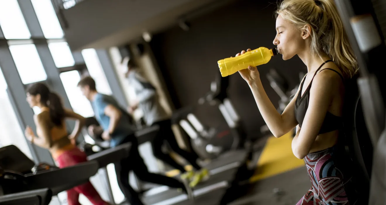 Mujer en un gimnasio tomando agua de una botella amarilla mientras otros usuarios corren en cintas de correr al fondo. La imagen muestra un ambiente moderno y activo, ideal para la fitness y el entrenamiento.