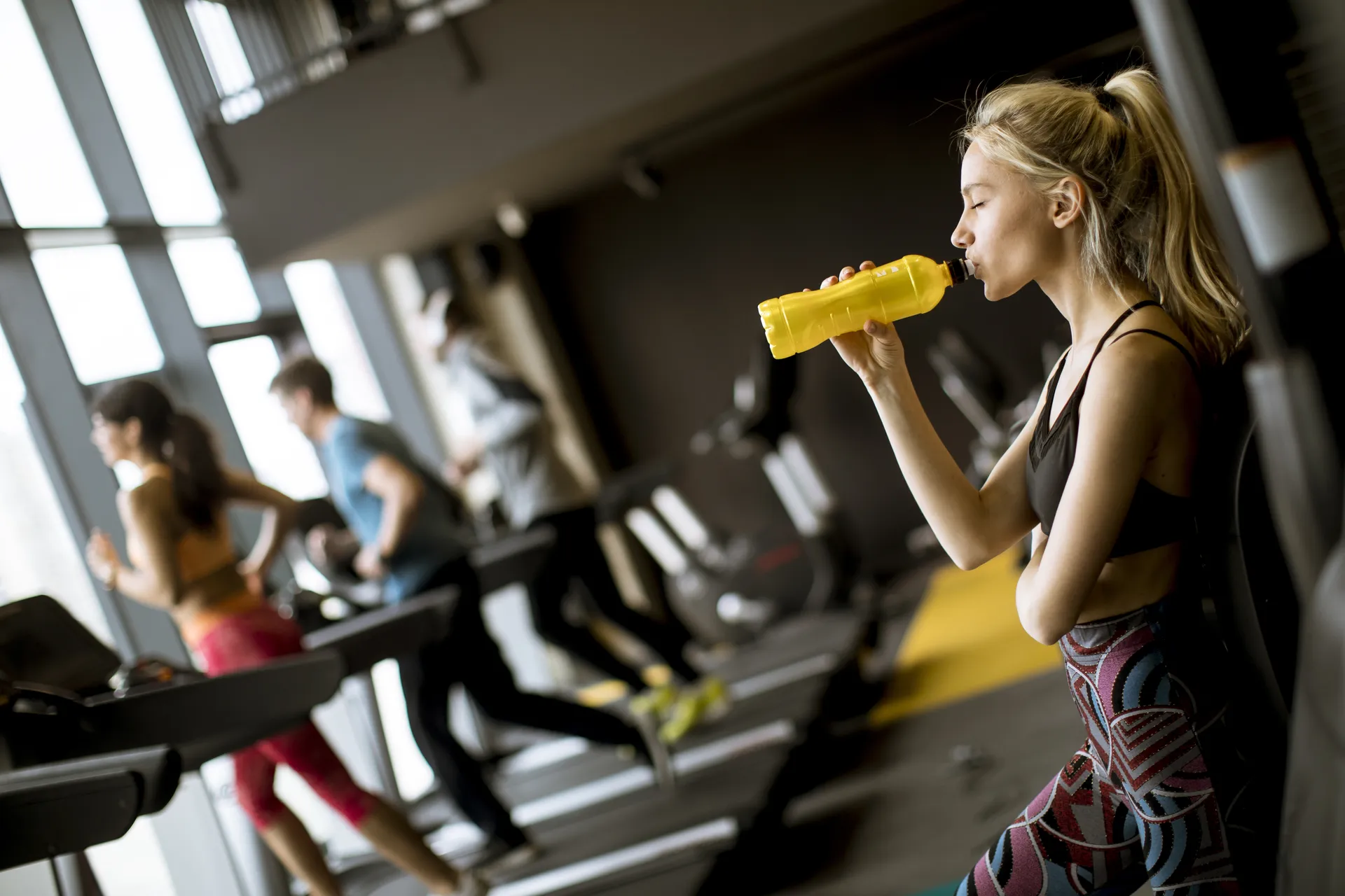 Mujer en un gimnasio tomando agua de una botella amarilla mientras otros usuarios corren en cintas de correr al fondo. La imagen muestra un ambiente moderno y activo, ideal para la fitness y el entrenamiento.